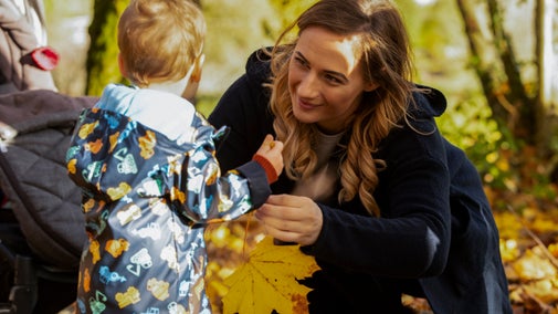 A woman and a child stand together in a wooded area, surrounded by colorful autumn leaves on the ground.